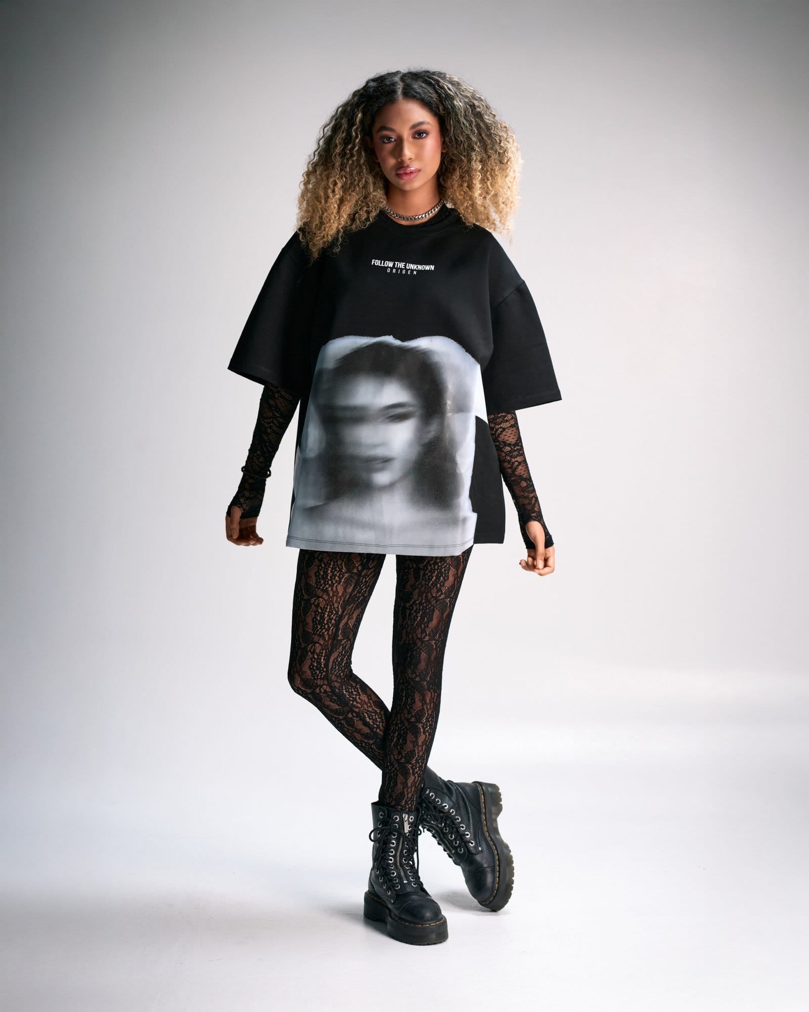 Bold fashion-forward young woman wearing an oversized black graphic t-shirt, lace gloves, patterned leggings, and chunky combat boots, showcasing edgy street style and modern urban fashion.