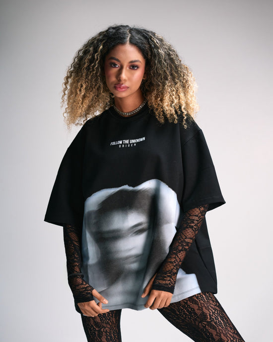 Bold young woman wearing a black oversized T-shirt with an artistic grayscale face print, layered with lace long sleeves and patterned leggings, showcasing edgy streetwear fashion.