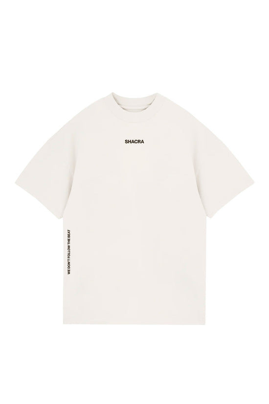 White oversized T-shirt with black branding "SHACRA" on the chest and side text "WE DON'T FOLLOW THE BEAT" printed vertically on the left sleeve, designed for streetwear fashion.