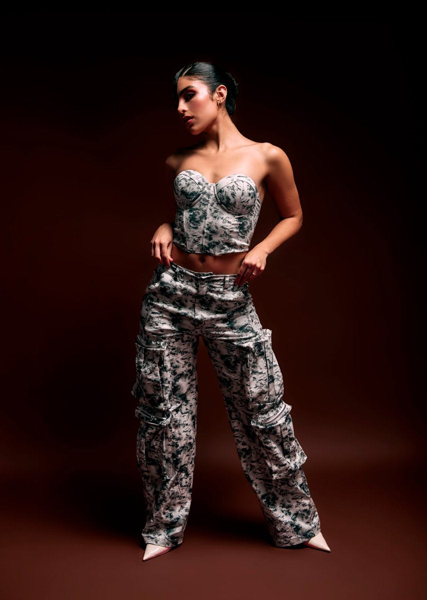 Stylish woman in camouflage-patterned crop top and cargo pants, black hair, bold makeup, posing confidently against dark brown background for fashion photography.